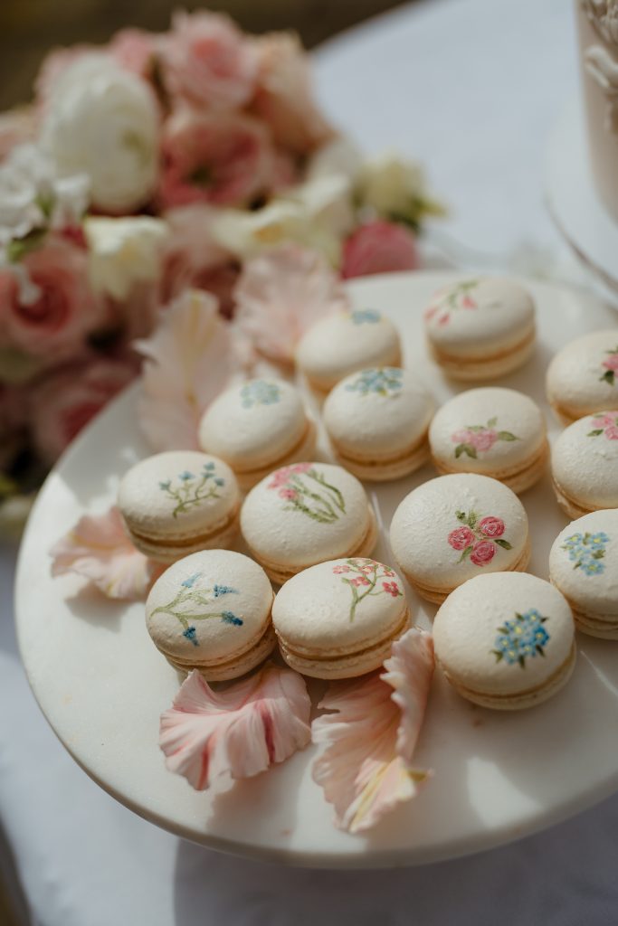 Elegant white macarons on a plate, each individually hand-painted with delicate pink and blue floral designs, suitable for luxury wedding favours or dessert tables.