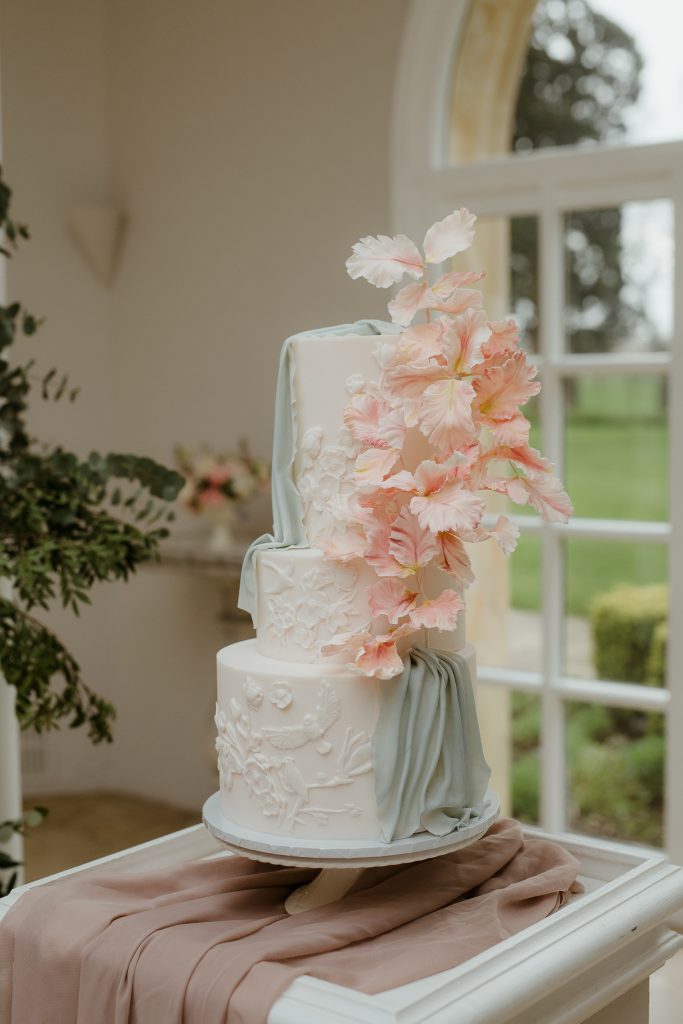 Elegant three-tiered white wedding cake with a sculptural design, featuring a cascade of delicate pink sugar flowers and a soft sage green edible drape, set in a bright, luxurious venue.