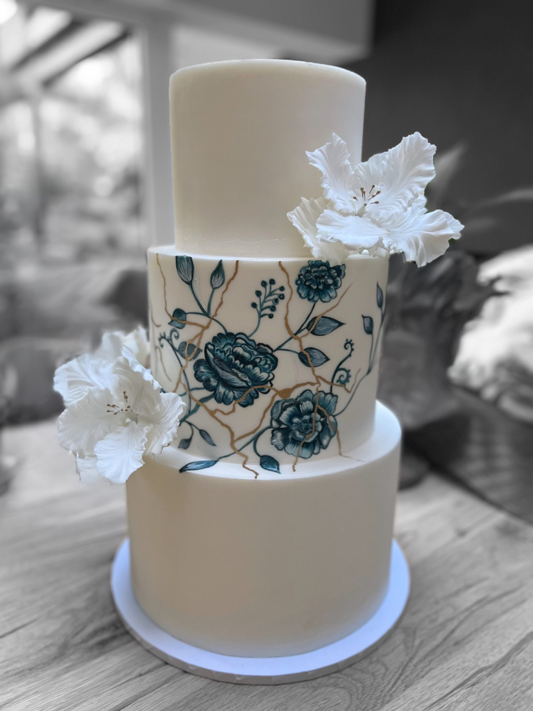 Elegant multi-tiered white wedding cake with a central tier featuring bespoke hand-painted blue and gold floral details, representing a personal story.