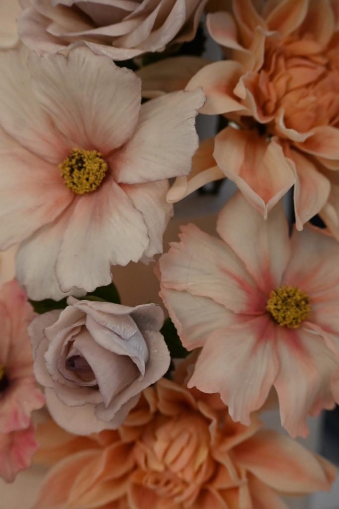 Close-up of incredibly lifelike handmade sugar flowers in shades of blush pink and peach, featuring detailed petals of roses, dahlias, and open blossoms.