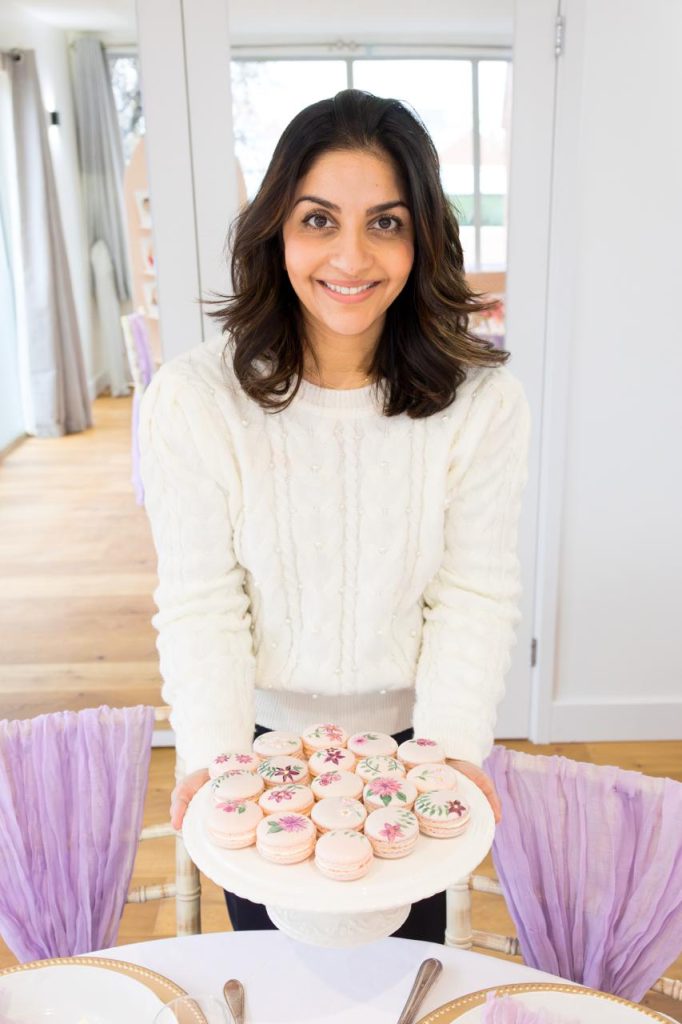 Dimple from Sugar Creations, smiling and holding a plate of elegant hand-painted floral macarons, representing bespoke luxury wedding confections and friendly service.
