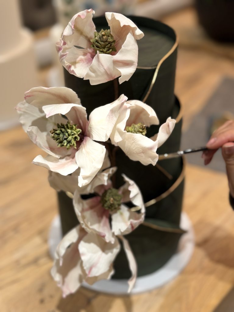 Modern luxury wedding cake in dark green with sculptural form, featuring large white sugar flowers and hand-painted gold detailing, showcasing 2025 trends.