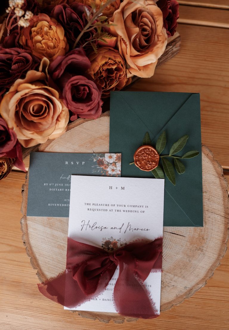 Elegant green and red wedding stationery displayed on a rustic log slice with seasonal flowers, styled for a Leicestershire wedding in the East Midlands.