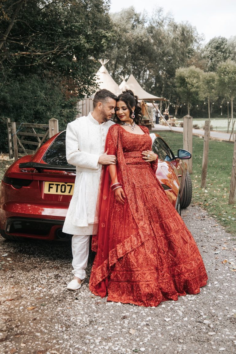 Bride and groom in traditional red Indian wedding attire posing beside a matching red car at The Hidden Hive wedding venue in the East Midlands.