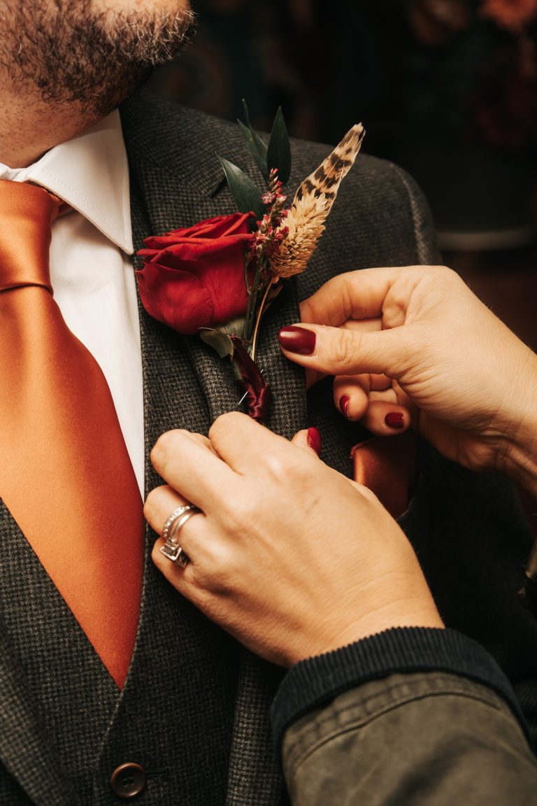 Close-up of a groom’s blazer as a red rose buttonhole is carefully pinned on before a Leicestershire wedding in the East Midlands.