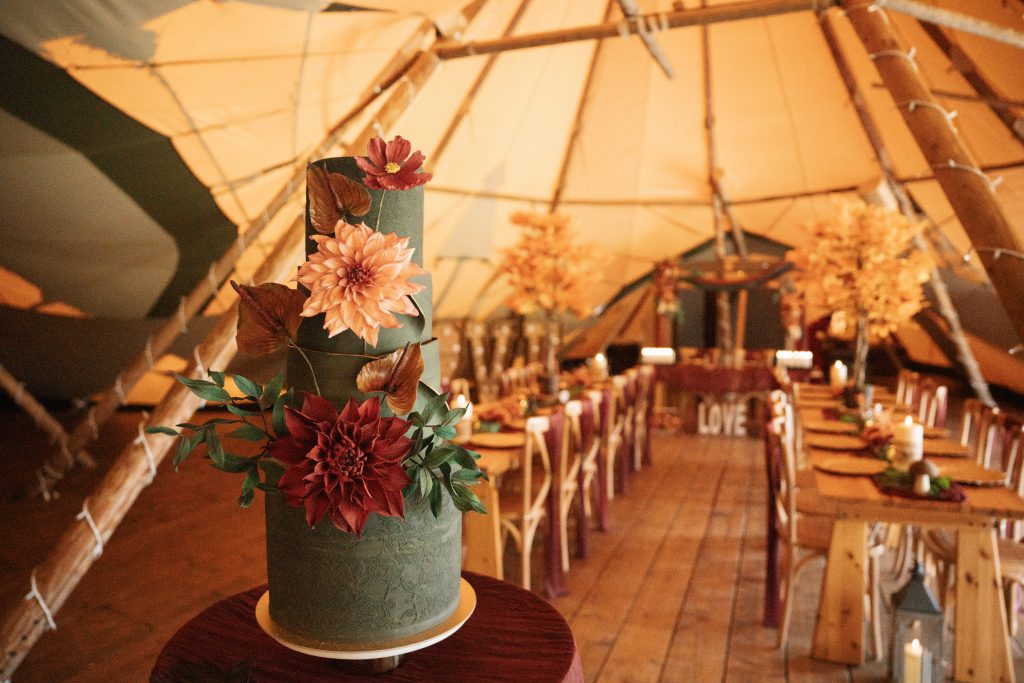Tall green wedding cake displayed inside a rustic tipi, decorated with warm autumn-inspired details and seasonal florals.
