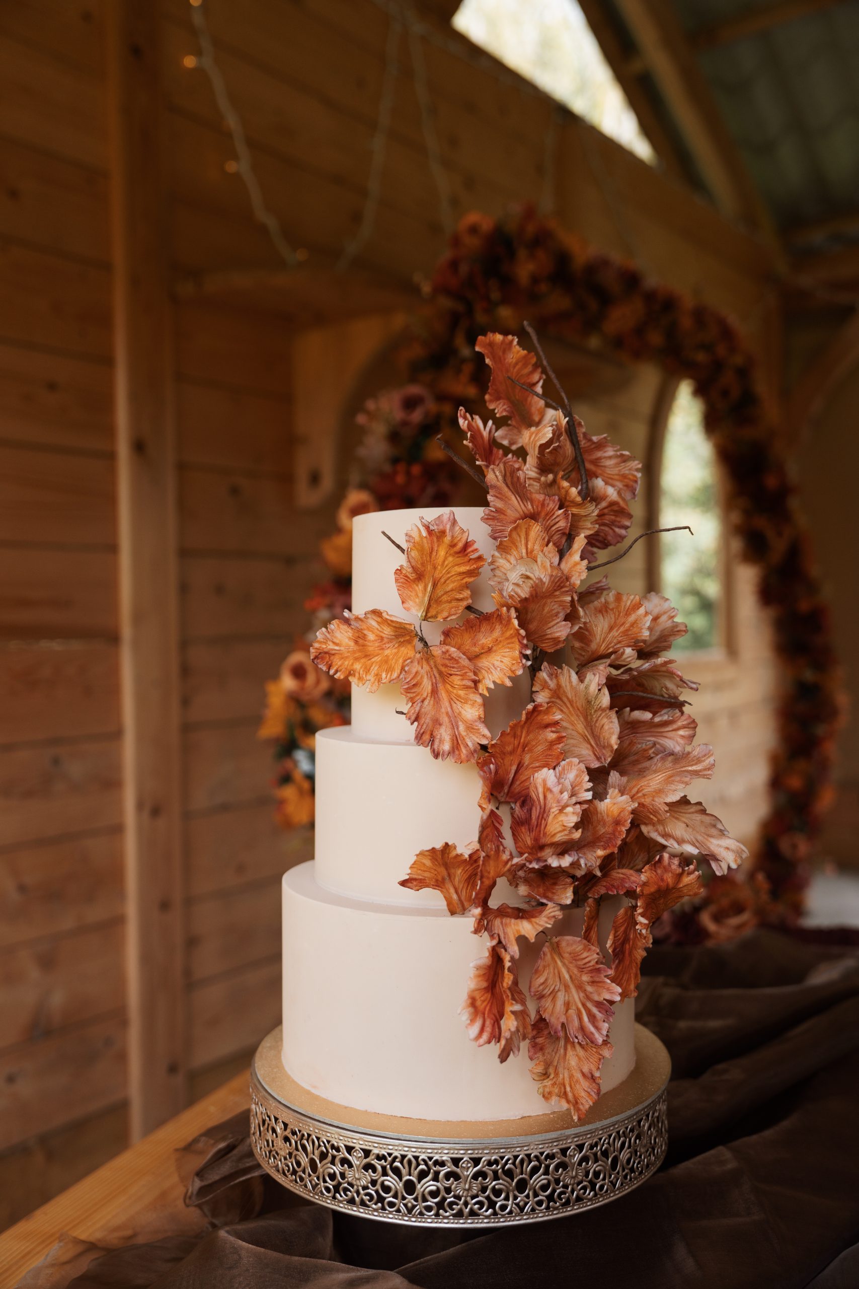 Nude three-tiered luxury wedding cake decorated with cascading burnt orange sugar leaves showing crisp autumn leaves on a gold stand.