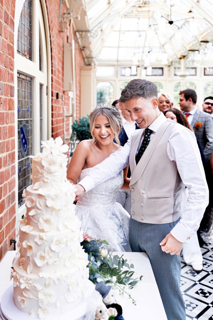 A joyful couple cutting a four-tier ivory wedding cake adorned with delicate handmade sugar flowers at Kilworth House in Leicestershire.