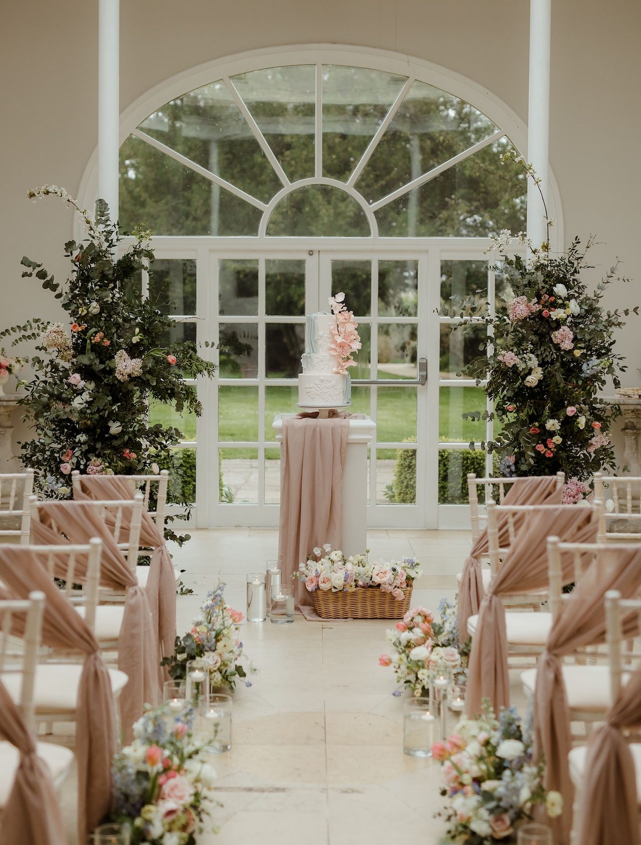 An elegant luxury wedding cake adorned with handmade sugar flowers in pastel pink and blue, displayed in the sunlit orangery at Stapleford Park with fresh florals surrounding it.