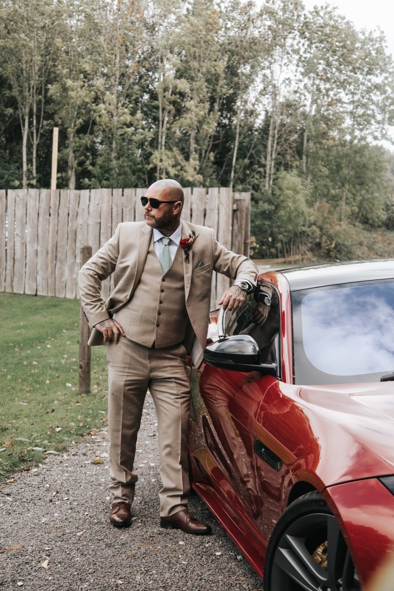 Groom wearing a beige suit posing confidently beside a red sports car at a stylish Leicestershire wedding in the East Midlands.