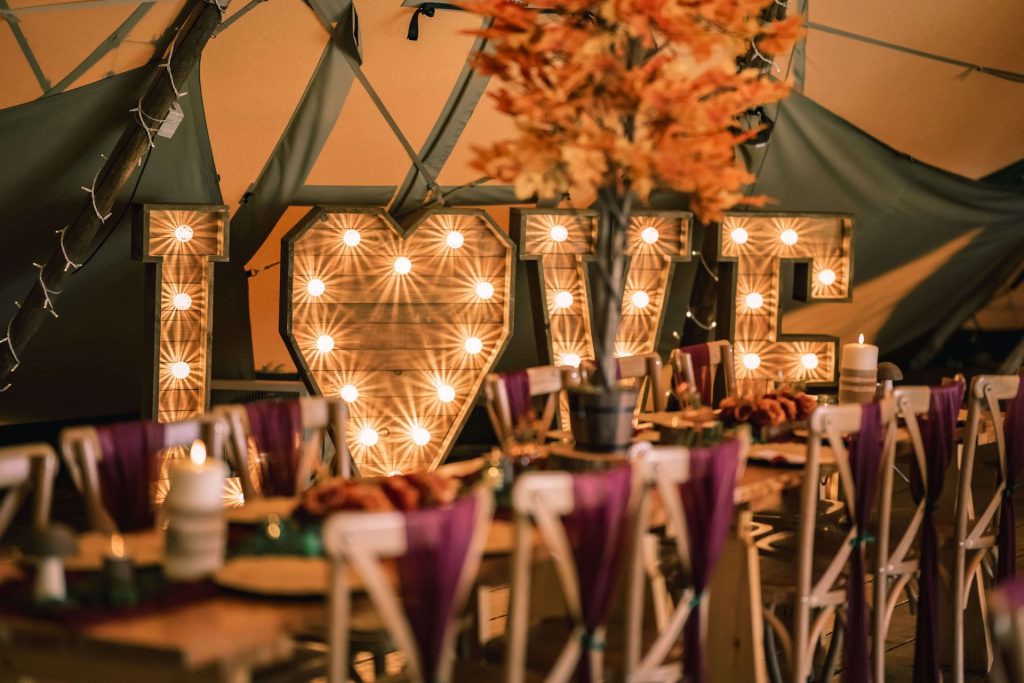 Warmly lit “LOVE” marquee letters glowing behind a rustic dining table decorated with candles, pumpkins, and autumn foliage.