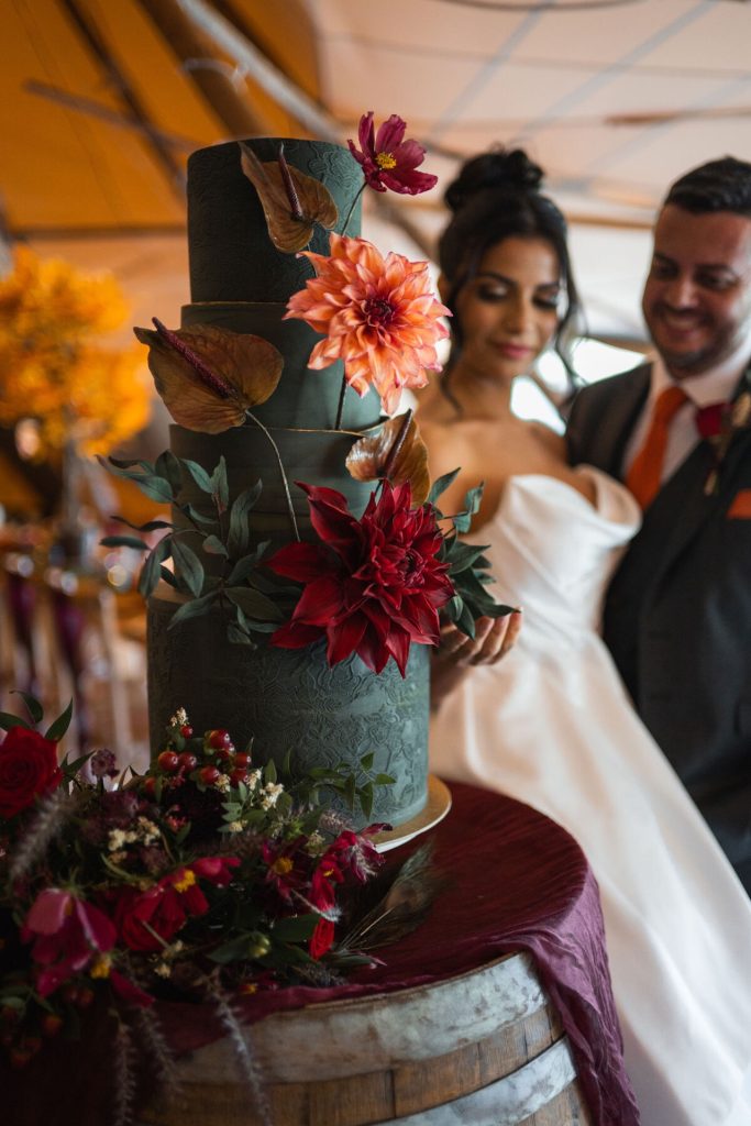 Green four-tier wedding cake decorated with handcrafted autumn sugar flowers, displayed on a rustic beer barrel as the bride and groom admire it.