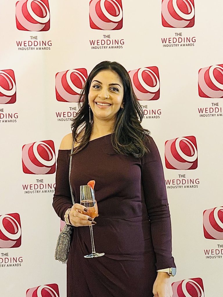Dimple dressed elegantly standing in front of The Wedding Industry Awards backdrop, holding a glass of champagne.