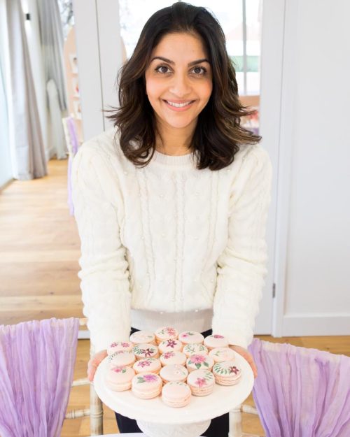 Dimple from Sugar Creations, smiling and holding a plate of elegant hand-painted floral macarons, representing bespoke luxury wedding confections and friendly service.